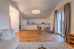 Een moderne woonkamer met een bank, een eettafel met stoelen, een keuken op de achtergrond en een groot raam met gordijnen.