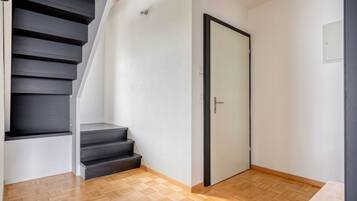 Een trap die leidt naar een kamer met een deur, houten vloeren en een lichtschakelaar aan de muur.