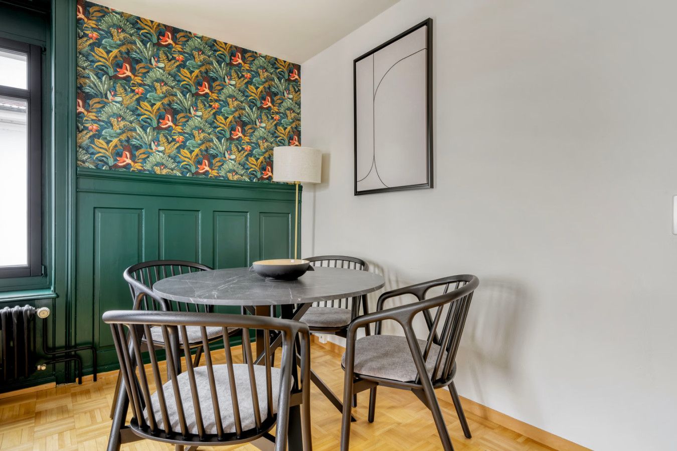 A dining area with a round table, four chairs, a decorative wall with a floral pattern, a green door, and a framed abstract artwork.