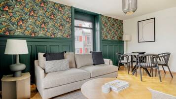 Een moderne woonkamer met een bank, een rond salontafeltje, een bijzettafeltje met een lamp, een eethoek met stoelen en groen behang met een patroon.