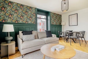 Een moderne woonkamer met een bank, een rond salontafeltje, een bijzettafeltje met een lamp, een eethoek met stoelen en groen behang met een patroon.