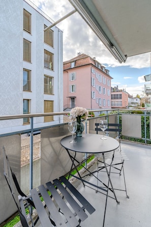 Een balkon met een ronde tafel, twee stoelen, een vaas met bloemen en een leuning.