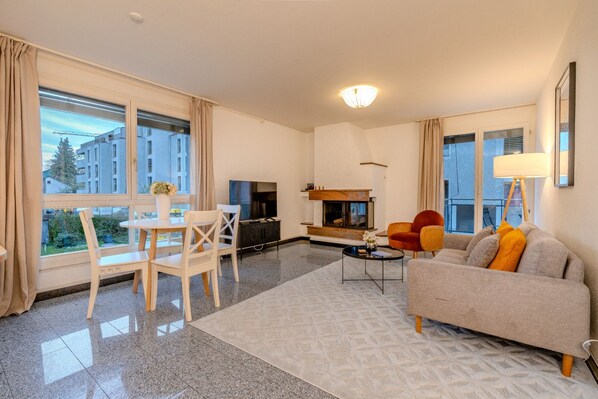 Een moderne woonkamer met een bank, stoelen, een salontafel, een open haard en grote ramen.