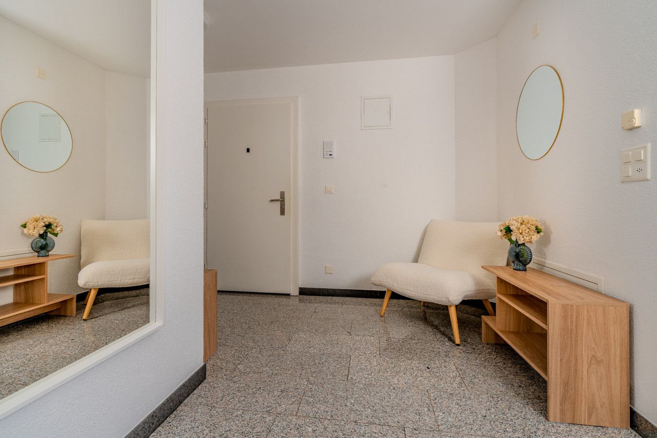 A hotel room with a large mirror, a beige armchair, a wooden console table, and a small wooden shelf.