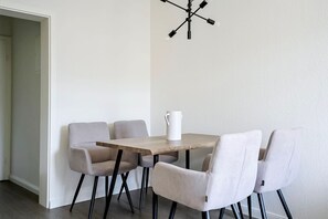 Uma sala de jantar com mesa de madeira e quatro cadeiras, um jarro branco sobre a mesa e uma luminária moderna na parede.