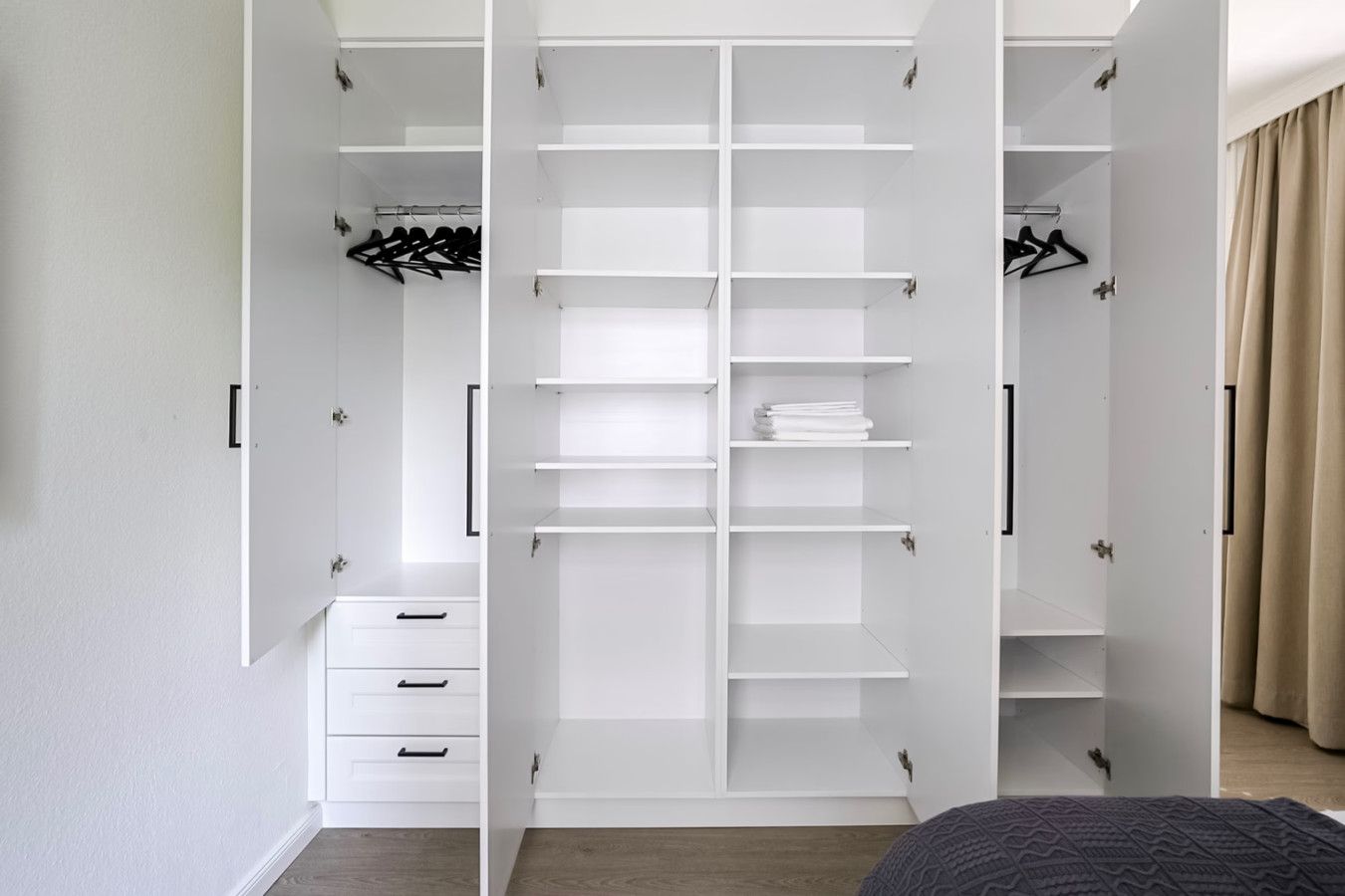 Ein weißer begehbarer Kleiderschrank mit mehreren Regalen und Platz zum Aufhängen, ein Bett mit dunkel gemustertem Bettbezug und ein beiger Vorhang auf der rechten Seite.