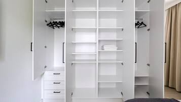 Ein weißer begehbarer Kleiderschrank mit mehreren Regalen und Platz zum Aufhängen, ein Bett mit dunkel gemustertem Bettbezug und ein beiger Vorhang auf der rechten Seite.