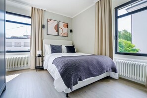 Quarto moderno com uma cama grande, mesa de cabeceira, janela com cortinas e arte abstrata na parede.