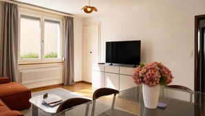 Ein modernes Wohnzimmer mit Fernseher, Esstisch aus Glas, einer Couch und einem Blumenstrauß.