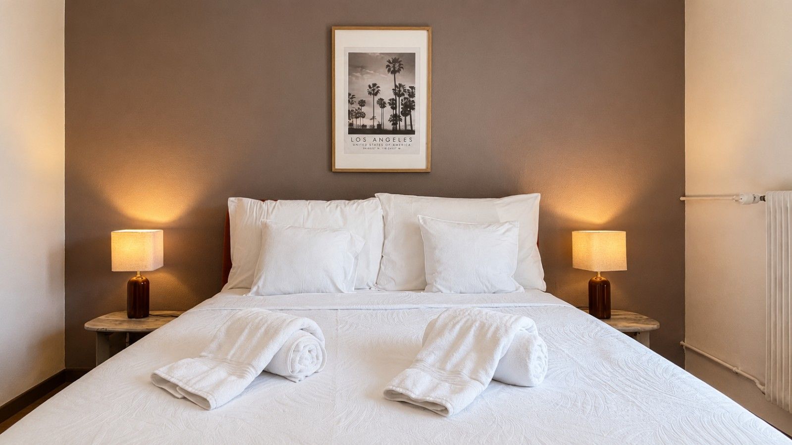 Une chambre d’hôtel avec un grand lit, deux lampes de chevet, un tableau encadré au mur et des serviettes roulées posées sur le lit.