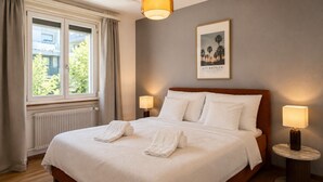 Ein ordentlich bezogenes Bett mit zwei aufgerollten Handtüchern, einem gerahmten Bild an der Wand und zwei Nachttischlampen.