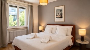 A neatly made bed with two rolled towels, a framed picture on the wall, and two bedside lamps. - Bright 2-BR in Wipkingen — ground floor, garden & tram nearby (Zürich)