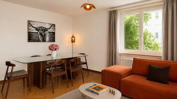 A modern living room with a glass dining table, wooden chairs, a sofa, a floor lamp, and a window with curtains.