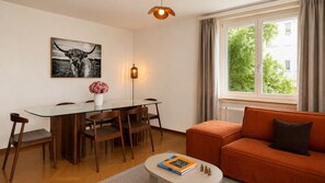 A modern living room with a glass dining table, wooden chairs, a sofa, a floor lamp, and a window with curtains. - Bright 2-BR in Wipkingen — ground floor, garden & tram nearby (Zürich)