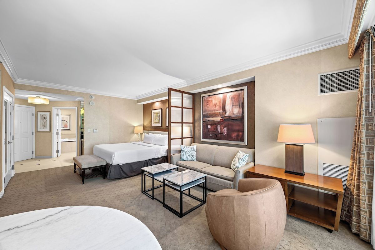 Ein Hotelzimmer mit einem Bett, zwei Nachttischen, einem Sofa, einem Couchtisch und einem großen Gemälde an der Wand.