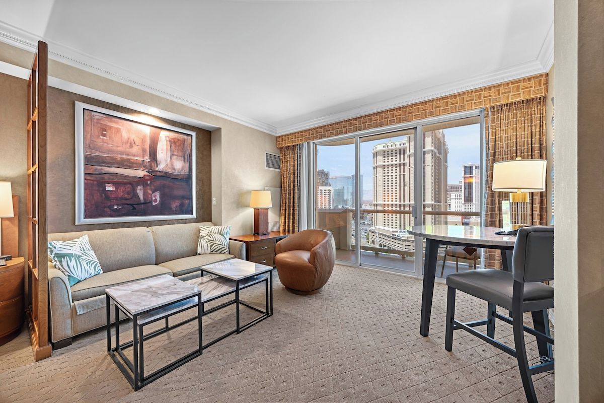 Habitación de hotel con sofá, sillón, mesa de centro y un ventanal que ofrece vistas a la ciudad.
