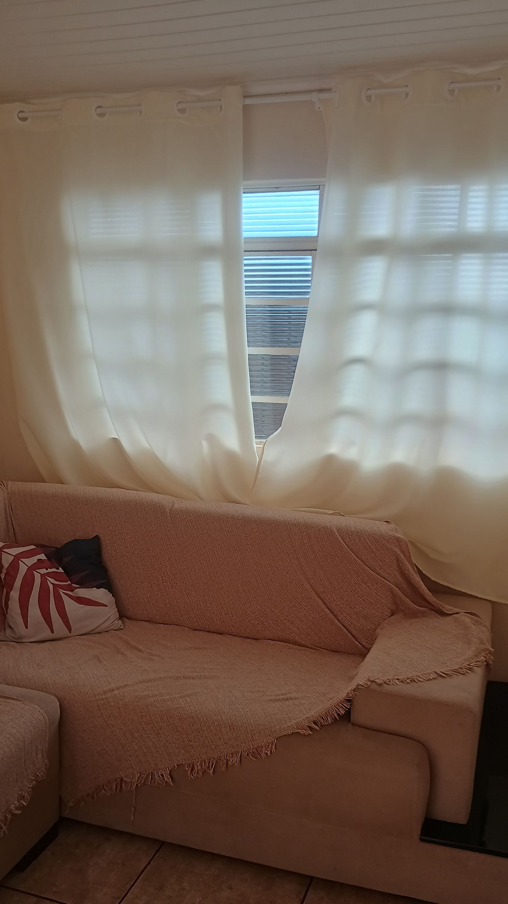 Ein Zimmer mit einem beigefarbenen Sofa, einem gemusterten Kissen und transparenten Vorhängen.