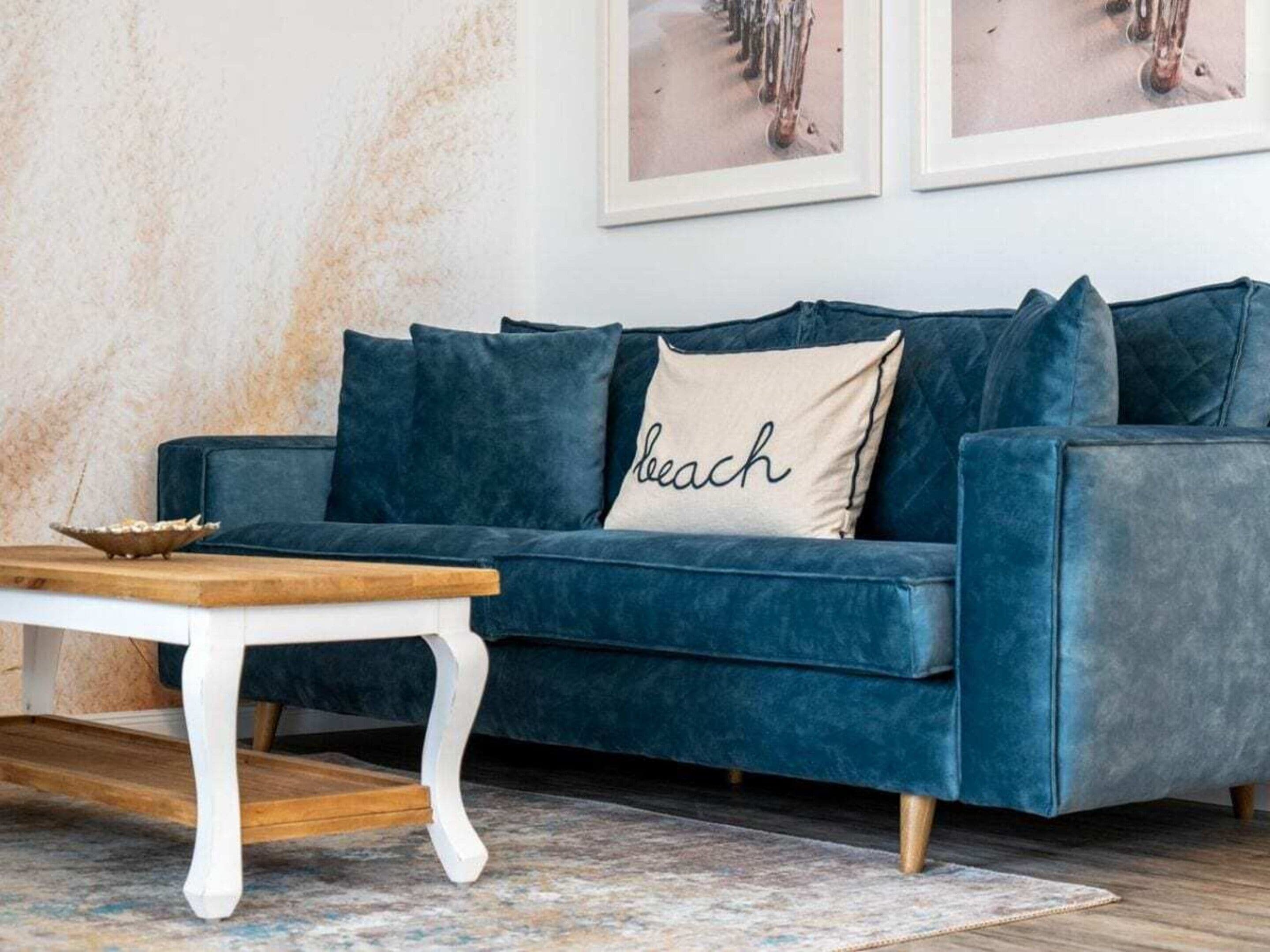 Een blauwe fluwelen bank met een kussen waarop 'beach' staat, een klein houten tafeltje en een kamerbreed tapijt met een patroon op de vloer.