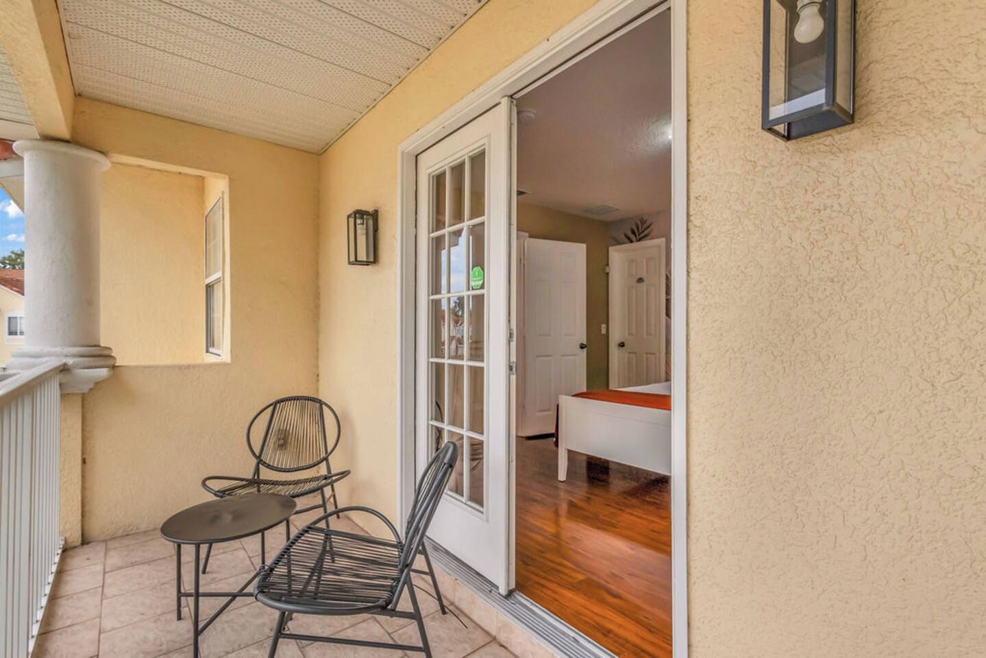Un balcon avec une table et deux chaises, une porte-fenêtre coulissante donnant sur une chambre, et un petit luminaire fixé au mur.