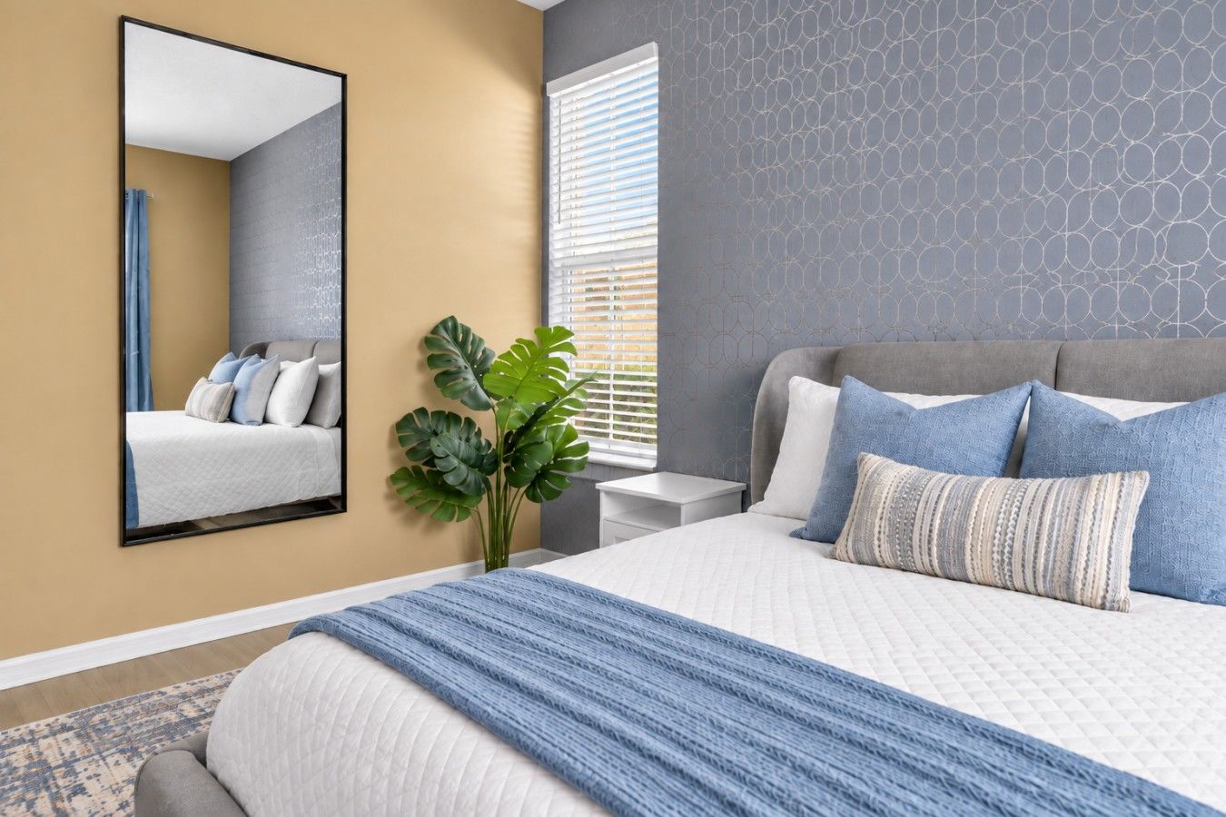 A bedroom with a bed, a nightstand, a mirror, and a window with blinds.