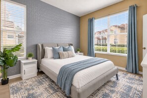 Quarto com cama, mesas de cabeceira, planta em vaso e vista de edifícios pela janela.