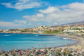 Une plage avec des parasols et des gens, un ciel dégagé et des bâtiments en arrière-plan.