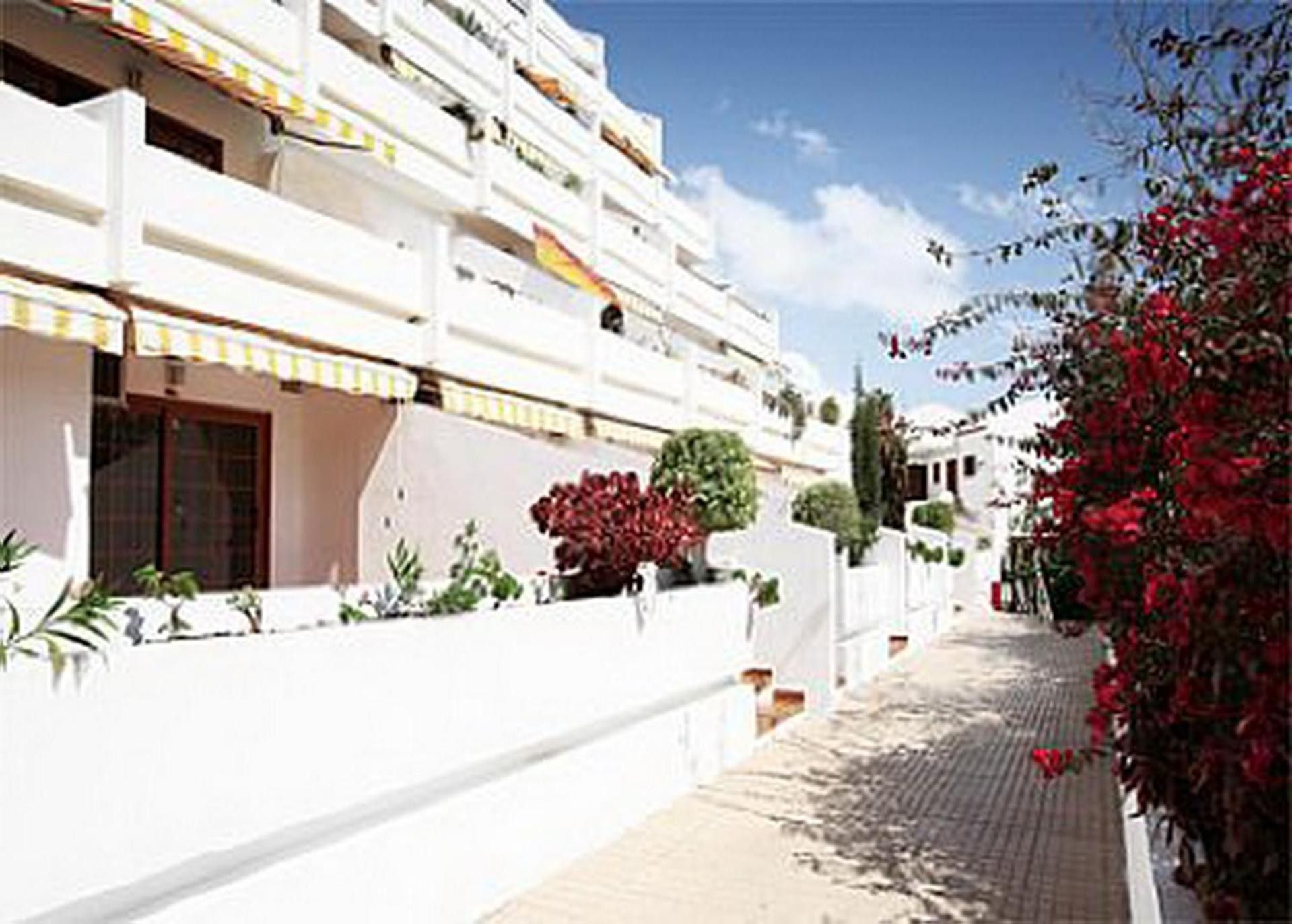 Un edificio blanco con varios balcones, un sendero pavimentado y plantas rojas con flores.