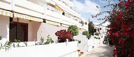 A white building with multiple balconies, a paved walkway, and red flowering plants.