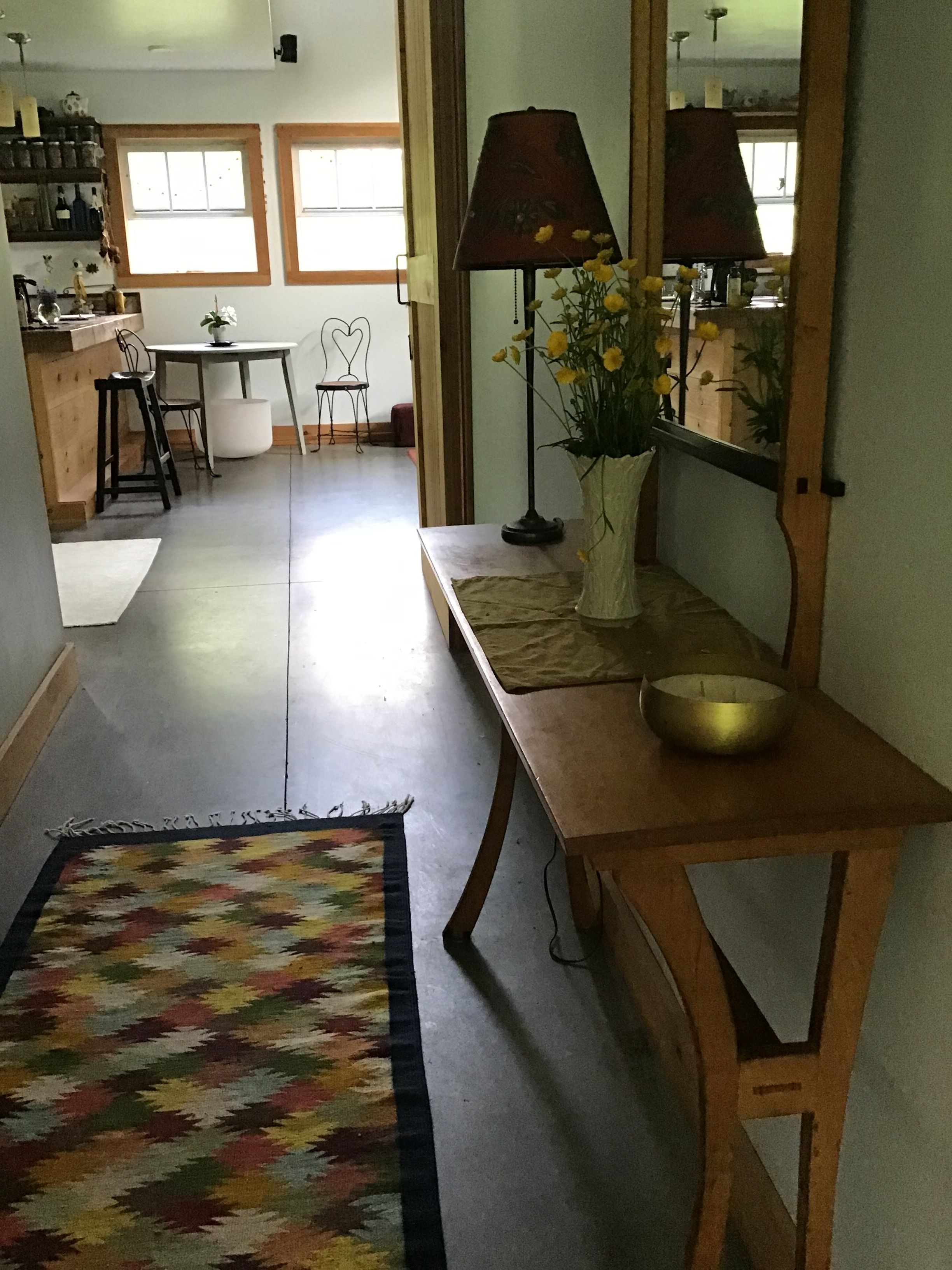 A hallway with a patterned rug, a wooden bench with a bowl, a lamp, and a mirror reflecting a room with a chair and a table.