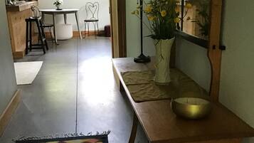 A hallway with a patterned rug, a wooden bench with a bowl, a lamp, and a mirror reflecting a room with a chair and a table.