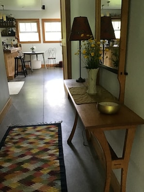 Un pasillo con un tapete estampado, un banco de madera con un cuenco, una lámpara y un espejo que refleja una habitación con una silla y una mesa.
