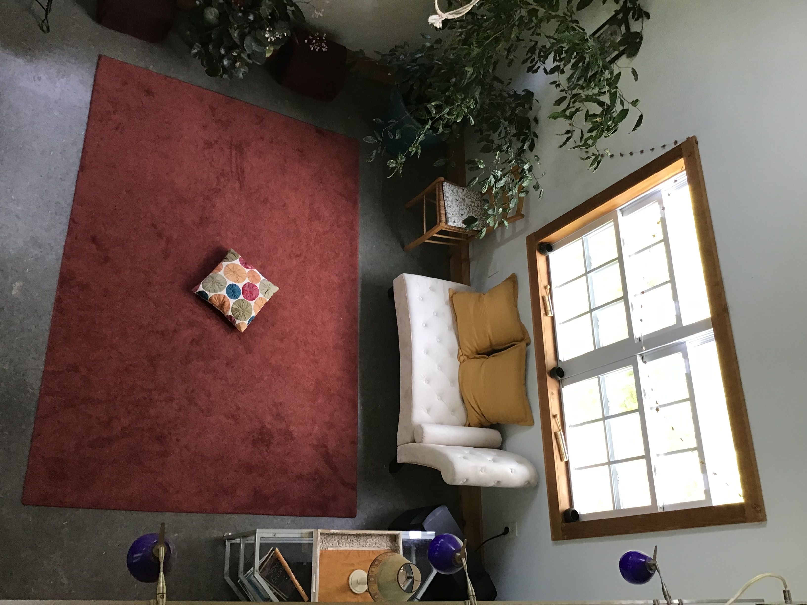 Une pièce avec une moquette rouge, un canapé blanc orné d’un coussin couleur moutarde, une petite table avec une lampe et une fenêtre avec des stores.