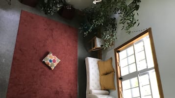 Une pièce avec une moquette rouge, un canapé blanc orné d’un coussin couleur moutarde, une petite table avec une lampe et une fenêtre avec des stores.