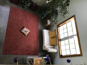 Una habitación con alfombra roja, un sofá blanco con un cojín mostaza, una mesita con una lámpara y una ventana con persianas.