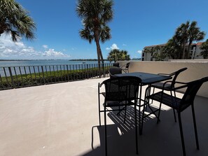 Eine Terrasse im Freien mit Tisch und Stühlen, mit Blick auf ein Gewässer und Palmen.