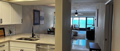 A modern kitchen with white cabinets, a stainless steel dishwasher, and a large window overlooking a balcony with outdoor furniture.