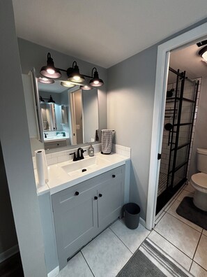 Ein Badezimmer mit einem weißen Waschbecken, einem schwarzen Wasserhahn und einem Spiegel darüber. Ein Handtuch hängt an der Wand und ein Mülleimer steht in der Ecke.