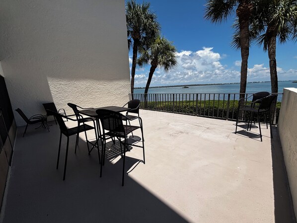 Eine Terrasse mit Tisch und Stühlen, Palmen und Blick aufs Wasser.