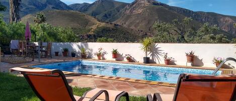 A swimming pool with chairs and potted plants, set against a mountainous backdrop.