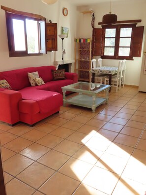 Ein Wohnzimmer mit einem roten Sofa, einem Glastisch, einem Esstisch mit Stühlen und einer Küchenzeile mit Holzschränken.