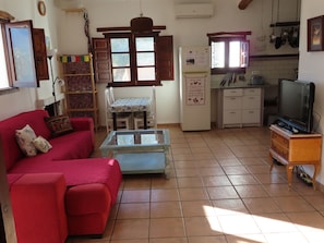 Ein Wohnzimmer mit einem roten Sofa, einem Glastisch, einem Fernseher und einer Küchenzeile inklusive Kühlschrank und Fenster.