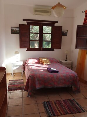 Un dormitorio con cama, mesita de noche, lámpara, aire acondicionado, ventana con contraventanas y vista a zonas verdes.