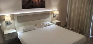 Een hotelkamer met een groot bed, twee nachtkastjes met lampen, een hoofdeinde en een raam met gordijnen.