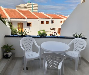Een balkon met een witte tafel en twee witte stoelen, planten in potten en uitzicht op gebouwen met rode dakpannen.