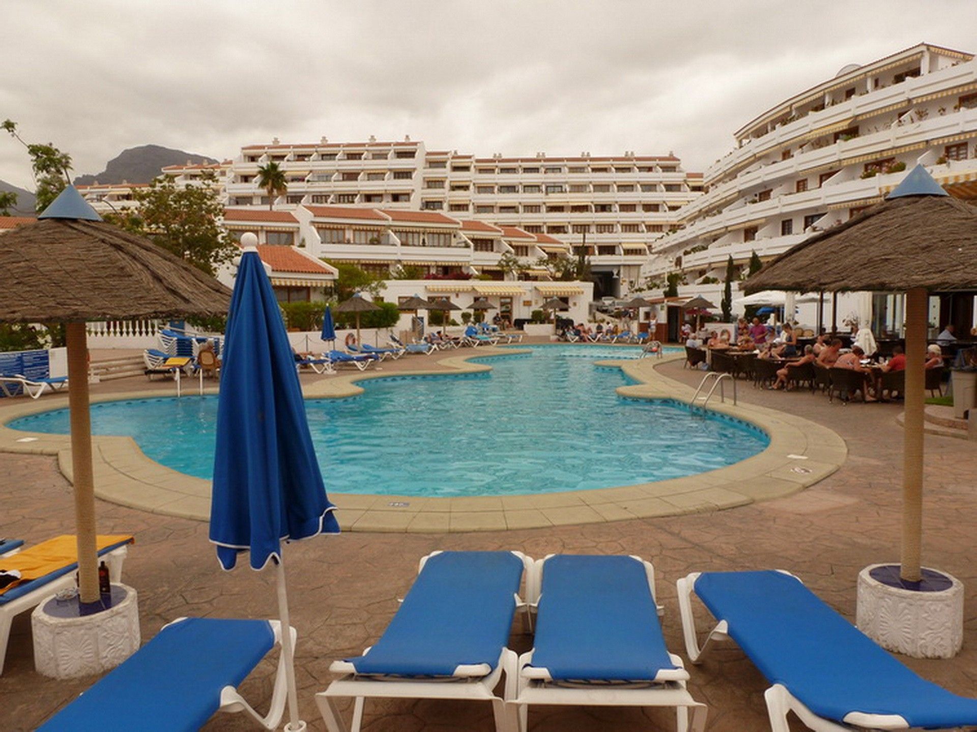 A hotel pool area with multiple lounge chairs and umbrellas, surrounded by a multi-story building with balconies.