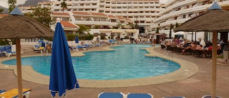 A hotel pool area with multiple lounge chairs and umbrellas, surrounded by a multi-story building with balconies.