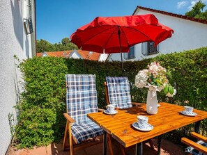 Un patio al aire libre con una mesa de madera, dos sillas a cuadros, un paraguas rojo y un florero con flores.
