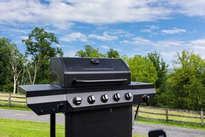 A black outdoor grill with a digital display and multiple control knobs, set against a backdrop of a grassy area and trees.