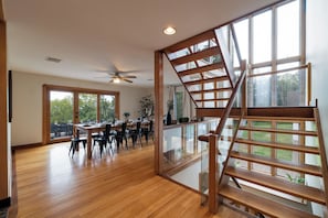 A spacious dining area with a wooden table and chairs, a staircase with glass railings, and large windows offering views of the outdoors.