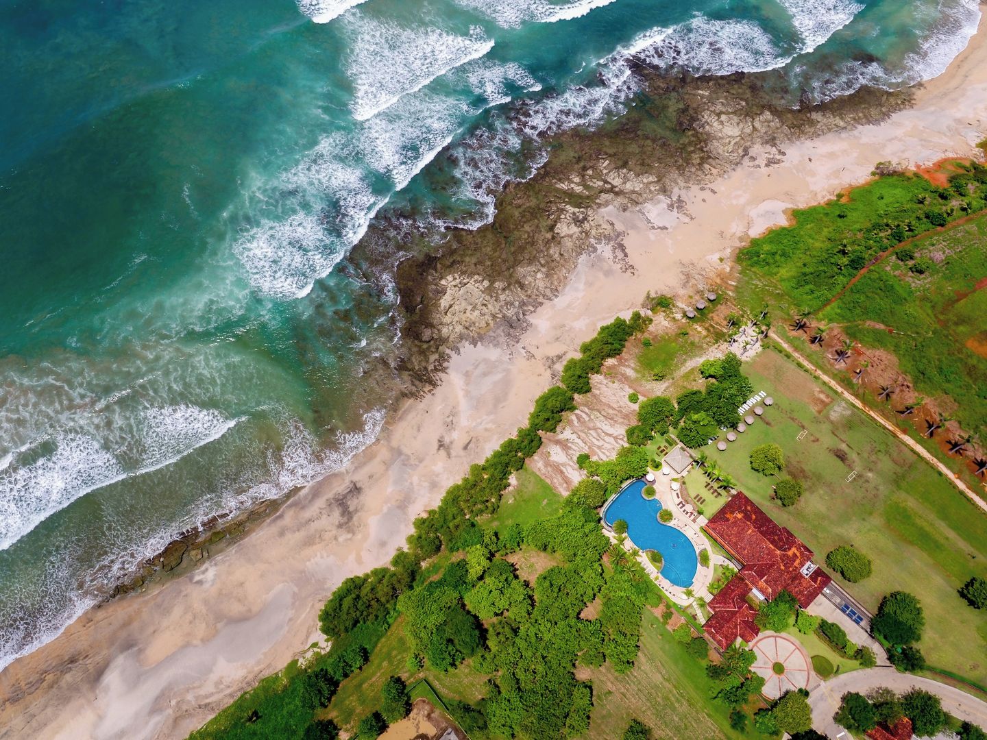Vue aérienne d’une propriété située sur la côte, avec une piscine, une végétation luxuriante et une plage.